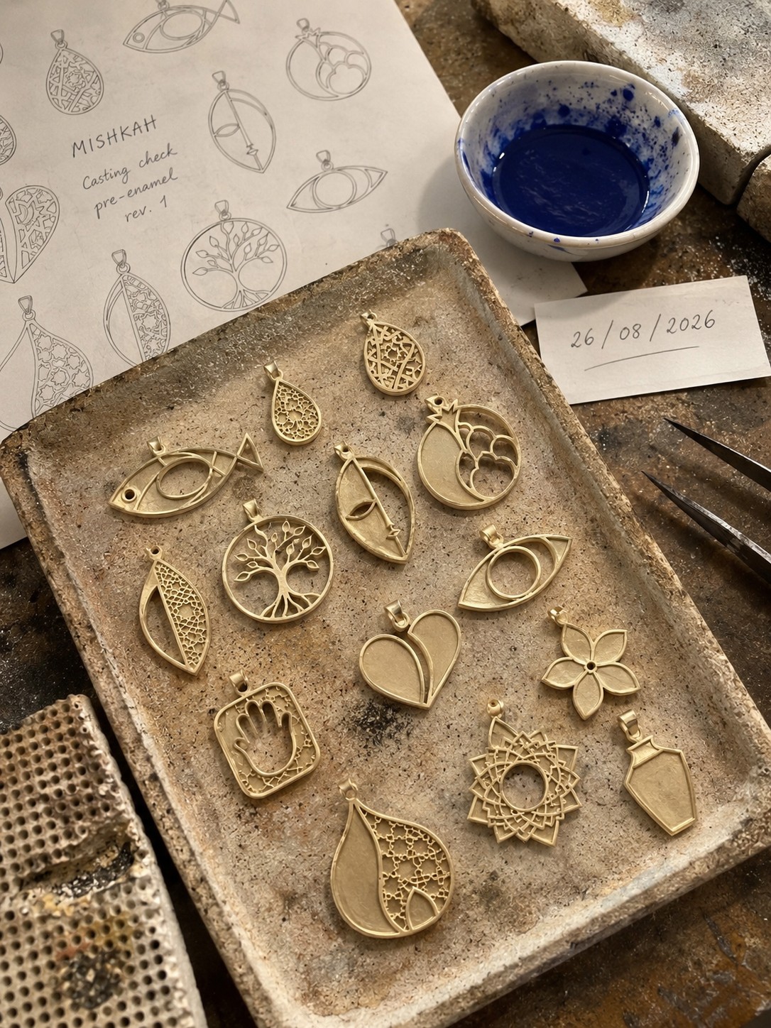 Twelve raw brass castings on a firing tray, before enamel, beside a dish of cobalt.